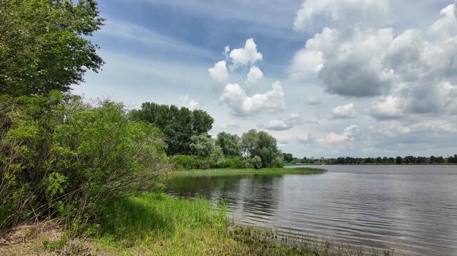 Scenic view of a calm lake with green trees on the far shore and dark grass in the foreground. Beautiful dramatic cloudy sky reflecting in the peaceful water during a summer day outdoors.
