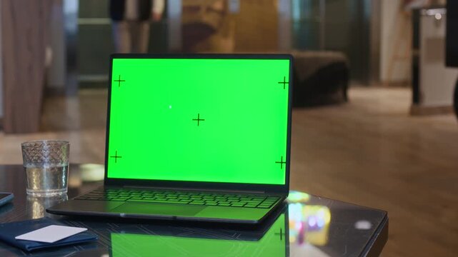 Laptop with green screen and tracking markers resting on glass table in modern hotel lobby while guests walking in blurred background. Ideal for inserting custom websites, Close Up