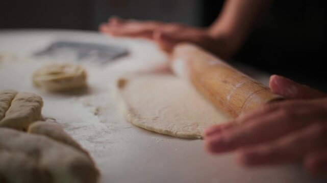 Hands roll dough on floured surface. Fingers guide wooden roller with care. Dough stretches thin as flour dusts air. Focus remains on skilled motion, not face. Perfect for cooking tutorials