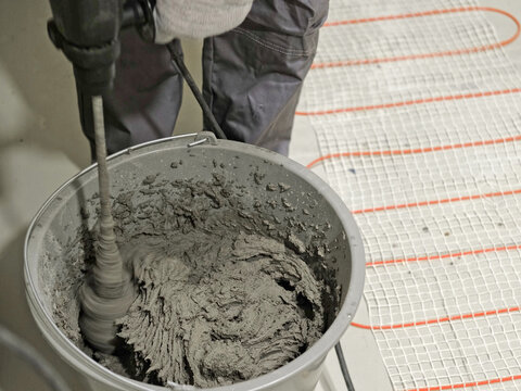 Construction worker preparing tile surface, mixing mortar substrate for flooring, underfloor heating mat. Workman combines adhesive in renovation setting. Operating power tool during bathroom upgrade