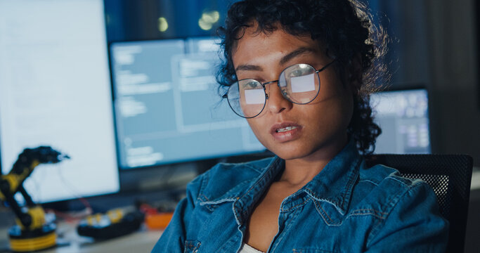 Close-up young Black female programmer coding AI software on multiple screens in night office. Network security, cybersecurity, machine learning, working late, overtime, robotics automation concept.