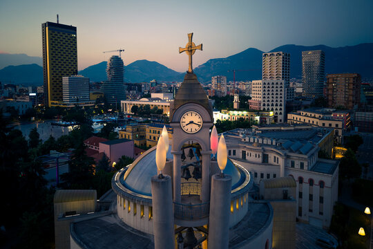 Aerial view of the Resurrection of Christ Orthodox Cathedral clock tower and the modern city skyline at dusk with mountains in the background in Tirane, Tirana County, Albania.