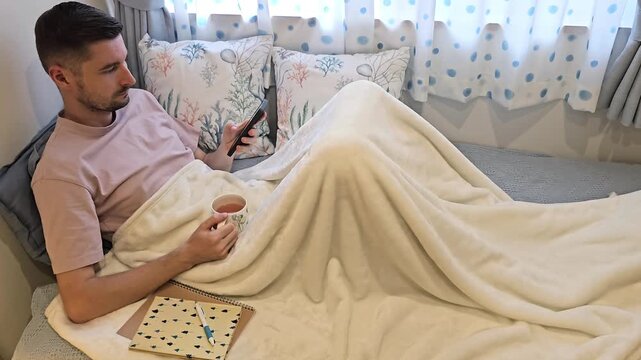 Young man scrolling social media feed on smartphone and drinking tea while relaxing in bed under blanket. Home lifestyle footage featuring mobile technology, leisure time and indoor comfort