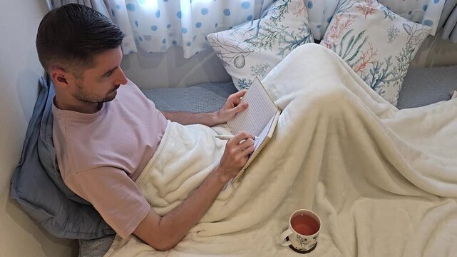 Young man sitting in bed under blanket writing in notebook with cup of tea nearby. Cozy home lifestyle scene with journaling, planning, studying and self reflection indoors. High angle view