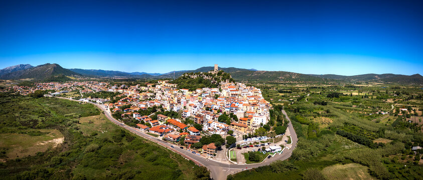 Aerial view of the historic town featuring the Castello della Fava on a hilltop surrounded by colorful houses and lush green plains under a clear blue sky in Posada, Sardinia, Italy.
