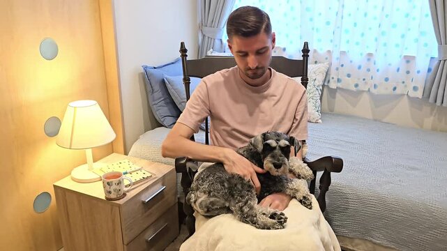 Young man sitting in rocking chair holding miniature schnauzer dog on lap in bedroom. Pet owner relaxing indoors with companion animal during quiet home leisure time