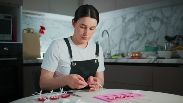 Young woman carefully crafts small decorations in modern kitchen. She wears black apron over white shirt. Focus is on her hands and detailed work. Kitchen has marble backsplash and soft lighting