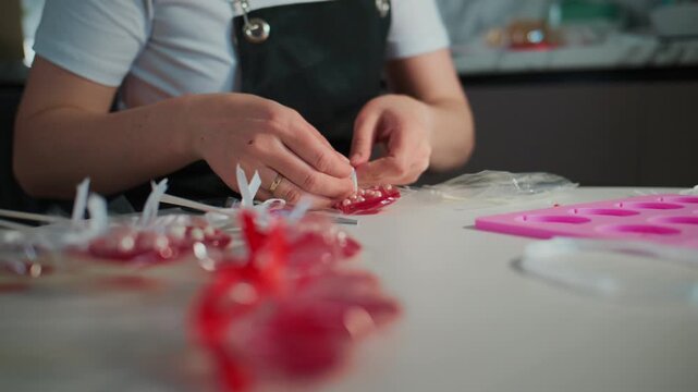 Person carefully assembles red candy hearts on a clean table. Hands focus intently on detailed crafting. Bright pink molds and wrapped candies surround the workspace