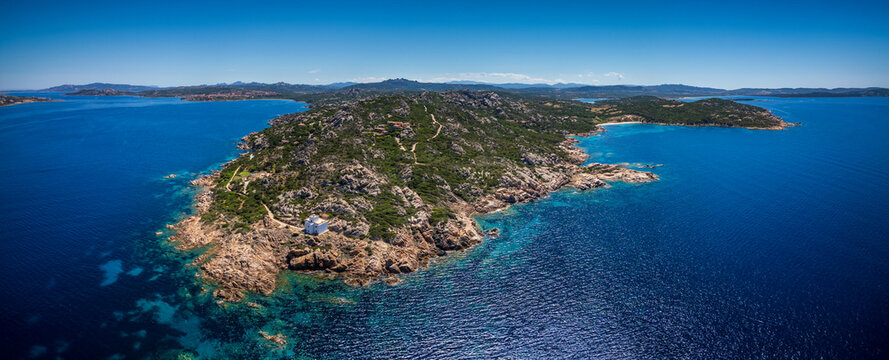 Aerial view of Faro di Punta Sardegna lighthouse on a rocky peninsula surrounded by turquoise Mediterranean waters and rugged hills in Palau, Sardegna, Italy.