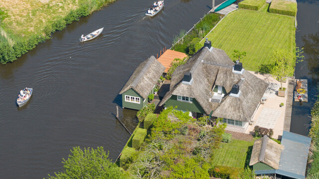 Aerial view of traditional thatched-roof houses along a canal with tourists in boats and lush green gardens in Giethoorn, Overijssel, Netherlands.