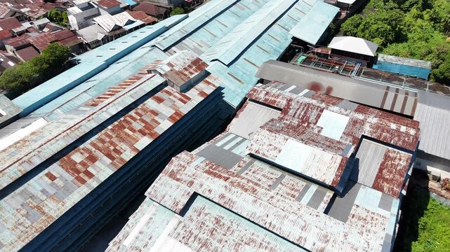 An aerial view of the weathered roofs of industrial warehouses with rusted corrugated metal sheets, along with the surrounding urban landscape.