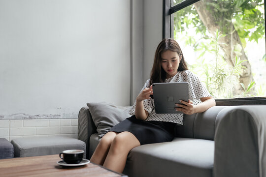 Young woman sitting on a sofa and using a tablet beside a window in a bright modern room, with a cup on the table and soft natural light