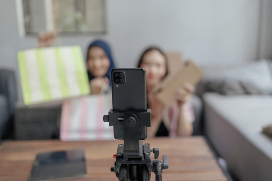 A smartphone mounted on a tripod records two blurred people holding colorful shopping bags in a bright living room with a sofa and coffee table in the background