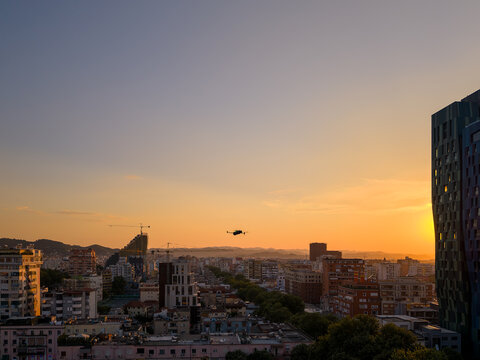Aerial view of urban skyline with modern buildings, construction cranes, and distant hills under a golden sunset sky with a drone flying in the foreground Tirana, Tirana County, Albania.