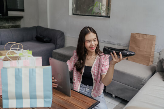 Young woman sitting on a sofa in a living room, smiling while holding a tablet and a shoe beside shopping bags and boxes in a casual home setting with natural light