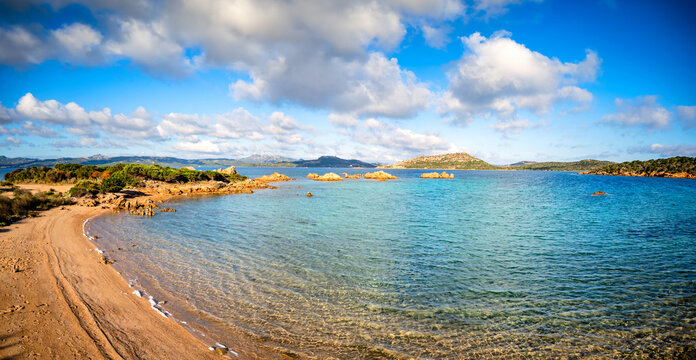 Aerial view of Spiaggia di Porto Palma with turquoise waters, golden sand, and rocky coastline under a blue sky with fluffy clouds La Maddalena, Sardegna, Italy.
