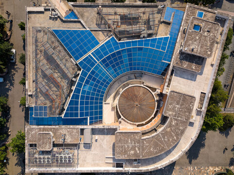Aerial view of the modern architecture of a building featuring a large blue glass roof and a circular central structure under bright sunlight in Tirane, Tirana County, Albania.