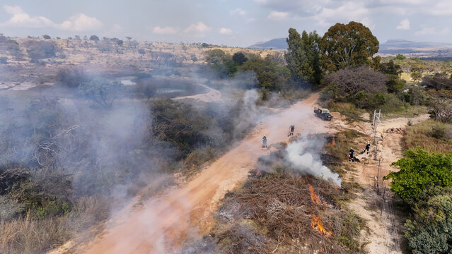 Aerial view of firefighters managing a controlled burn along a dirt road with smoke rising from dry brush and trees Vaalwater, Limpopo, South Africa.