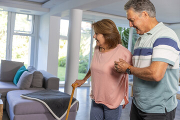 Obraz premium Mature man in striped polo steadying Senior woman in top holding wooden cane in living room