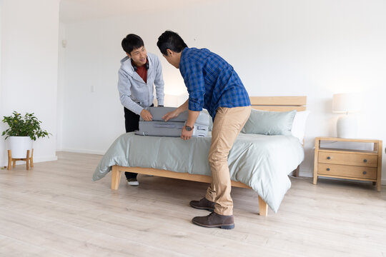 Asian men lifting and placing large gray hard-shell suitcase on bed in bright minimalist bedroom