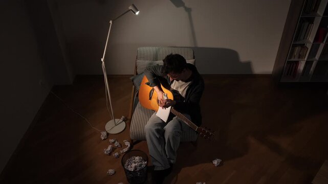 Man feeling frustrated while writing lyrics and composing music, surrounded by crumpled papers and a guitar in a dimly lit room, symbolizing the creative process and struggle