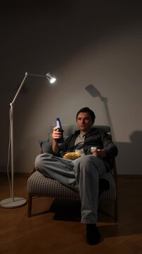 Vertical video man unwinding in an armchair, holding a beer bottle, remote control, and a bowl of chips under a soft lamp light, savoring a moment of evening leisure and entertainment at home