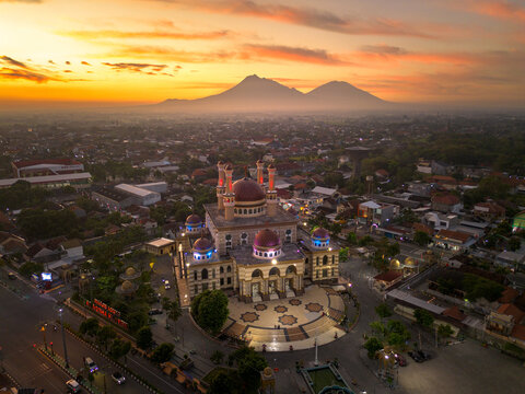Aerial view of the Great Mosque of Klaten with illuminated domes and minarets against a backdrop of volcanic peaks during a golden sunset Indonesia.