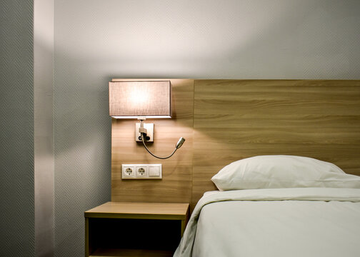 Modern hotel room interior with wooden headboard, wall mounted lamp, white bedding, bedside table, electrical outlets, minimalist design, contemporary accommodation.