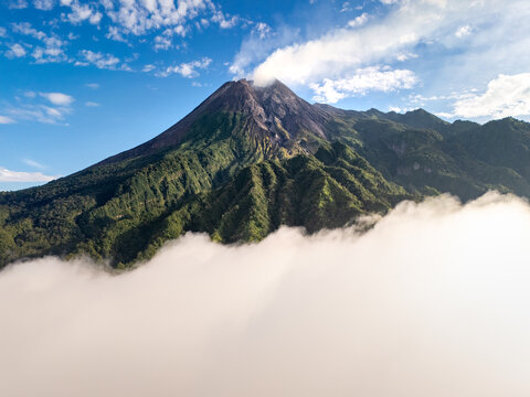 Aerial view of Mount Merapi volcano with smoke emitting from the crater above a thick layer of white clouds and lush green forested slopes Indonesia.