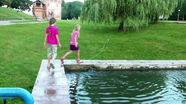 Two little sisters walk around a small city pool. Smolensk, Russia.