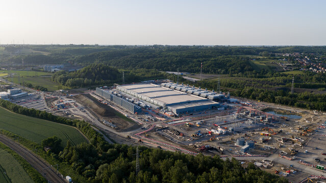 Aerial view of the Charleroi SAM 3A data center under construction with large industrial buildings and cooling units surrounded by green hills in Farciennes, Region wallonne, Belgium.