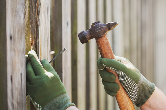 Male worker hands drive nail with hammer tool in rusrtic old wooden fence plank, maintenance repair, diy and carpentry outdoor.