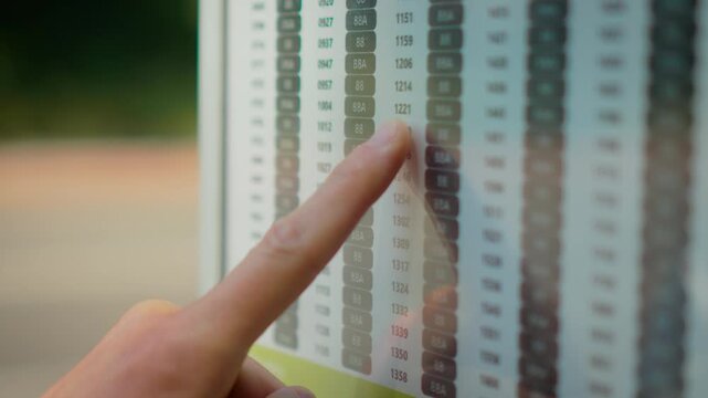 Checking bus timetable at public transport stop on empty urban street during daytime. Finger following schedule board with route numbers and departure times for city commuting. Public transportation