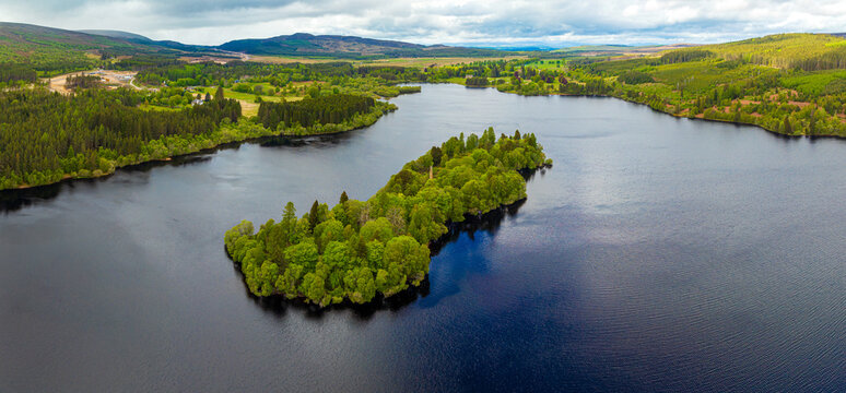 Aerial view of a lush green island in the middle of a calm lake surrounded by dense forests and rolling hills under a cloudy sky in Tomatin, Scotland, United Kingdom.