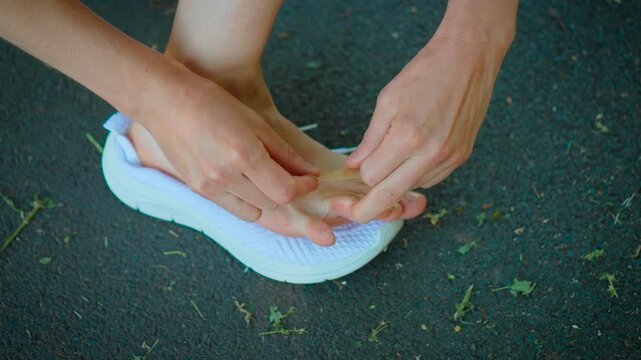 Applying adhesive bandage on blistered toe after walking or running outdoors. Closeup female hands covering foot skin irritation while wearing white athletic sneakers on asphalt path. Foot care