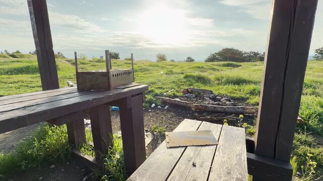 A rustic, wooden picnic setup sits abandoned in a sun-drenched, grassy field. The presence of litter and worn surfaces evokes a lonely, neglected feeling within otherwise beautiful natural landscape.
