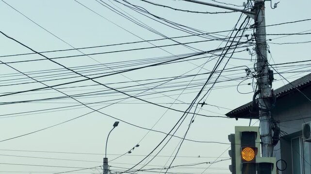 A chaotic mess of black electric wires and cables tangles across a pale evening sky from an old utility pole. The messy street infrastructure creates a stressful, overwhelming, and chaotic feeling.