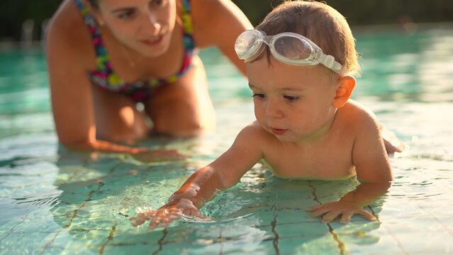 Cheerful young mom teaching her infant toddler how to float and swim in a bright pool during a summer day. Concept of early childhood development, water safety, and bonding time