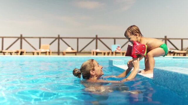 Mom in a bikini helps her son in oversleeves to jump from the side to the pool with clear water
