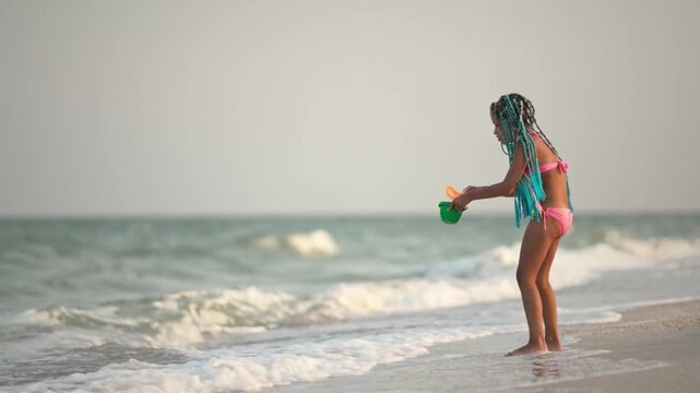 A girl with african braids in a summer costume plays on the beach with shells near the sea with waves under sunny sunset