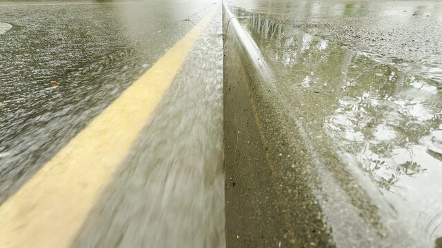Heavy rain floods a city roadway. Water streams rapidly down the paved asphalt gutter, producing a constant rushing and splashing sound.