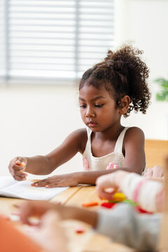Young African girl writes classwork while classmates handle colorful block during back to school primary classroom activity, notebook, pencil, concentration, study habit, responsibility, progress grow