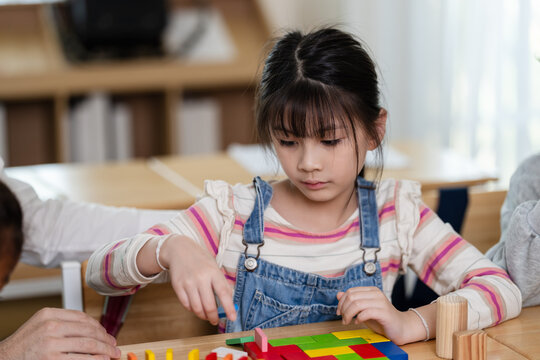 Asian elementary girl places colorful block during teacher guided desk activity, building pattern thinking, attention, classwork participation, back to school learning confidence, and early math skill