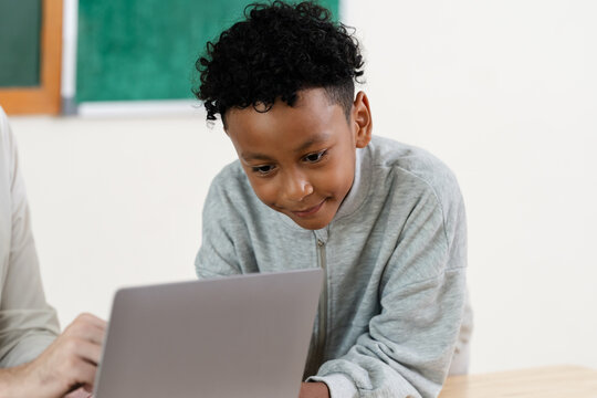 Elementary boy smiles while using laptop during teacher supported classroom task, building digital learning confidence, classwork routine, attention, problem solving, school readiness, and progress