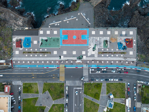 Aerial view of a colorful basketball court and playground situated on the rocky volcanic coastline with a busy road and green park in Catania, Sicily, Italy.