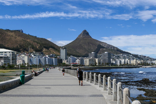 People walk along the Sea Point promenade on a sunny day in Cape Town, with Lion's Head in the distance