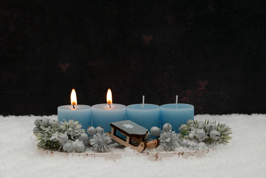 Second Sunday of Advent: An arrangement featuring two burning blue candles, a small wooden sled, and fir branches in the snow, set against a dark background with ample space for text.