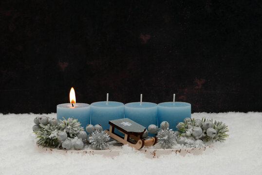 First Sunday of Advent: An arrangement featuring a burning blue candle, a small wooden sled, and fir sprigs in the snow, set against a dark background with ample space for text.