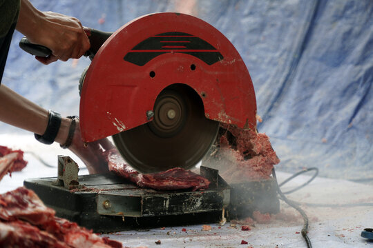 An electric bone saw machine chopping thick beef bones and raw ribs during Islamic Eid al Adha. Close-up of butcher hand slicing hard sacrificial animal bones for qurban meat division process.