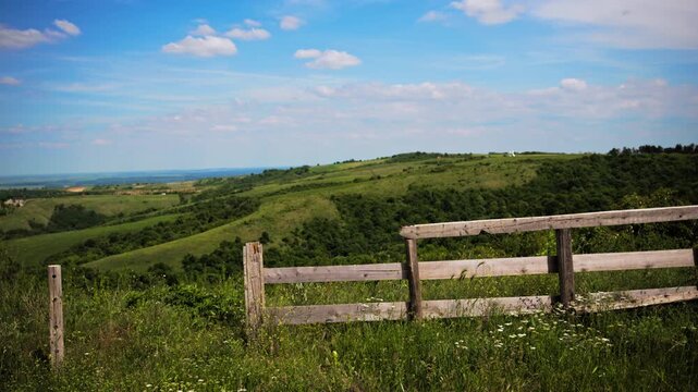 Rolling green hills with rustic wooden fence under blue sky and scattered clouds, peaceful rural countryside landscape, 4K.